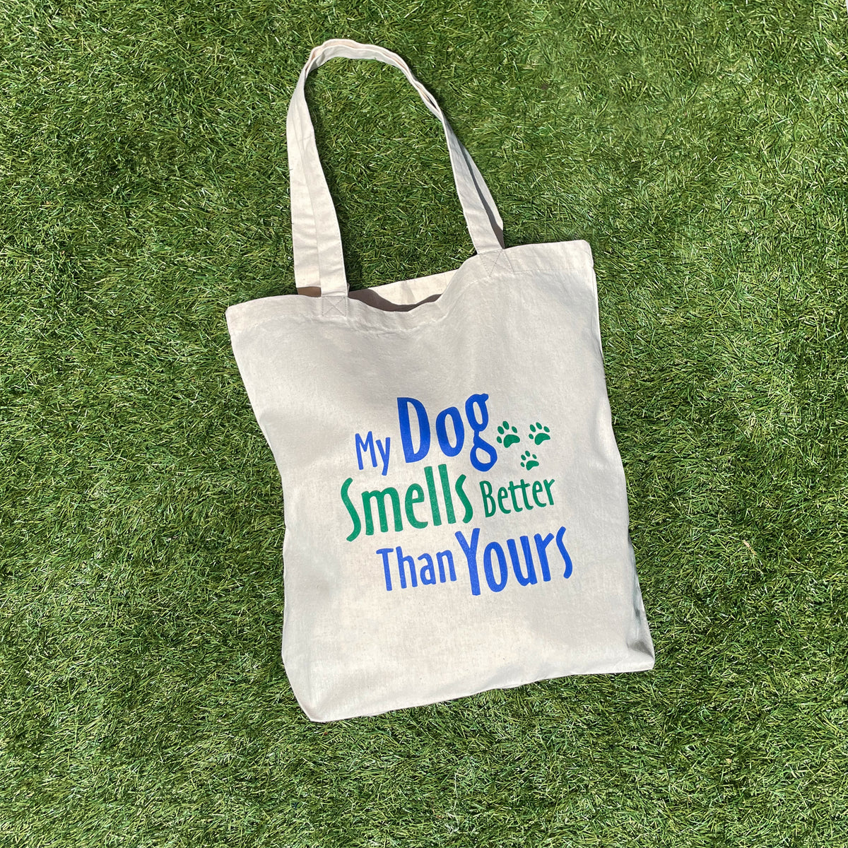 A beige reusable shopping bag with the text 'MY DOG SMELLS BETTER THAN YOURS' printed on the front on the grass.